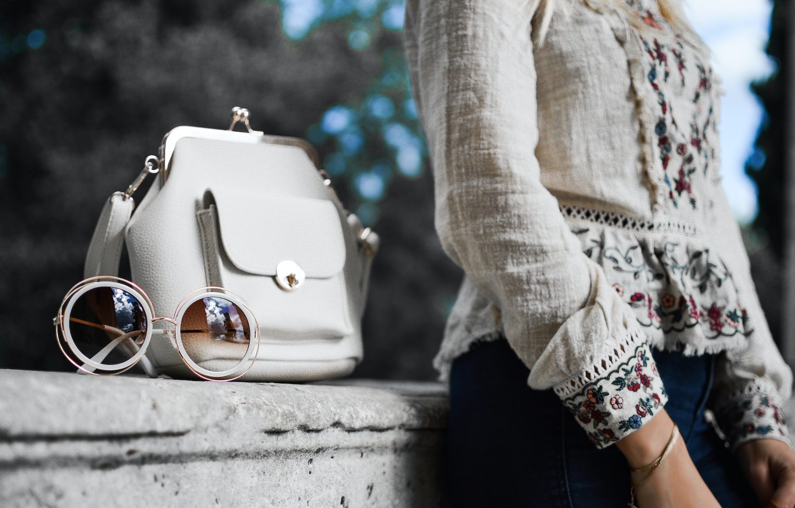 woman wearing beige and red floral top leaning on gray concrete slab with white leather bag ontop
