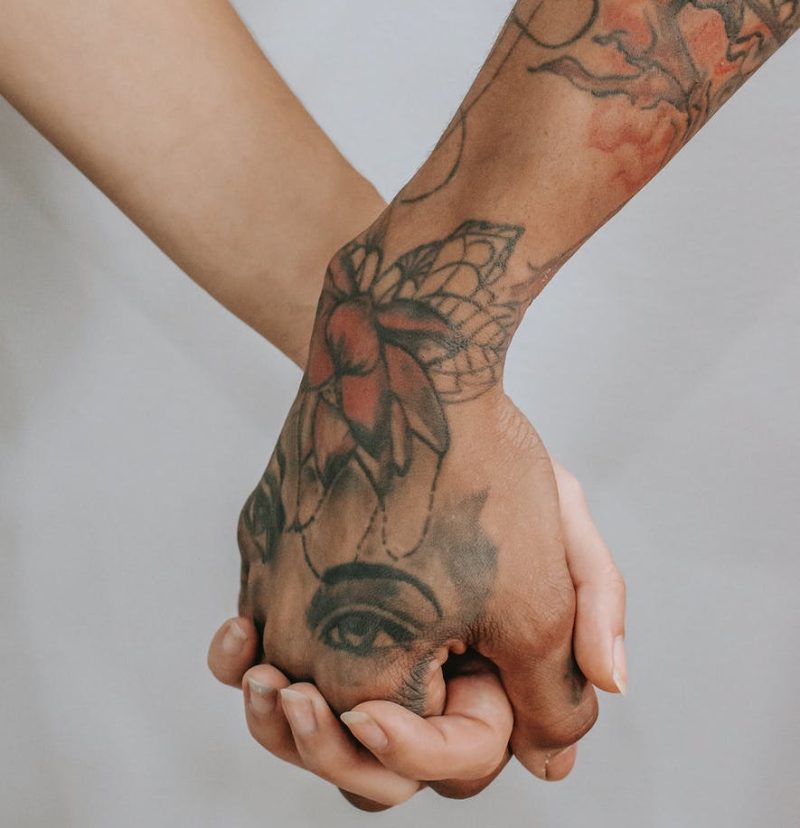 Young female hand holding other ethnic person hand with tattoos on gray background