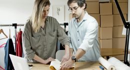 Two people preparing a package at a workspace with clothing racks and boxes in the background