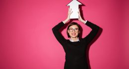 Smiling woman in glasses holding a white arrow sign above her head against a bright pink background