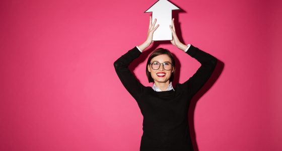 Smiling woman in glasses holding a white arrow sign above her head against a bright pink background